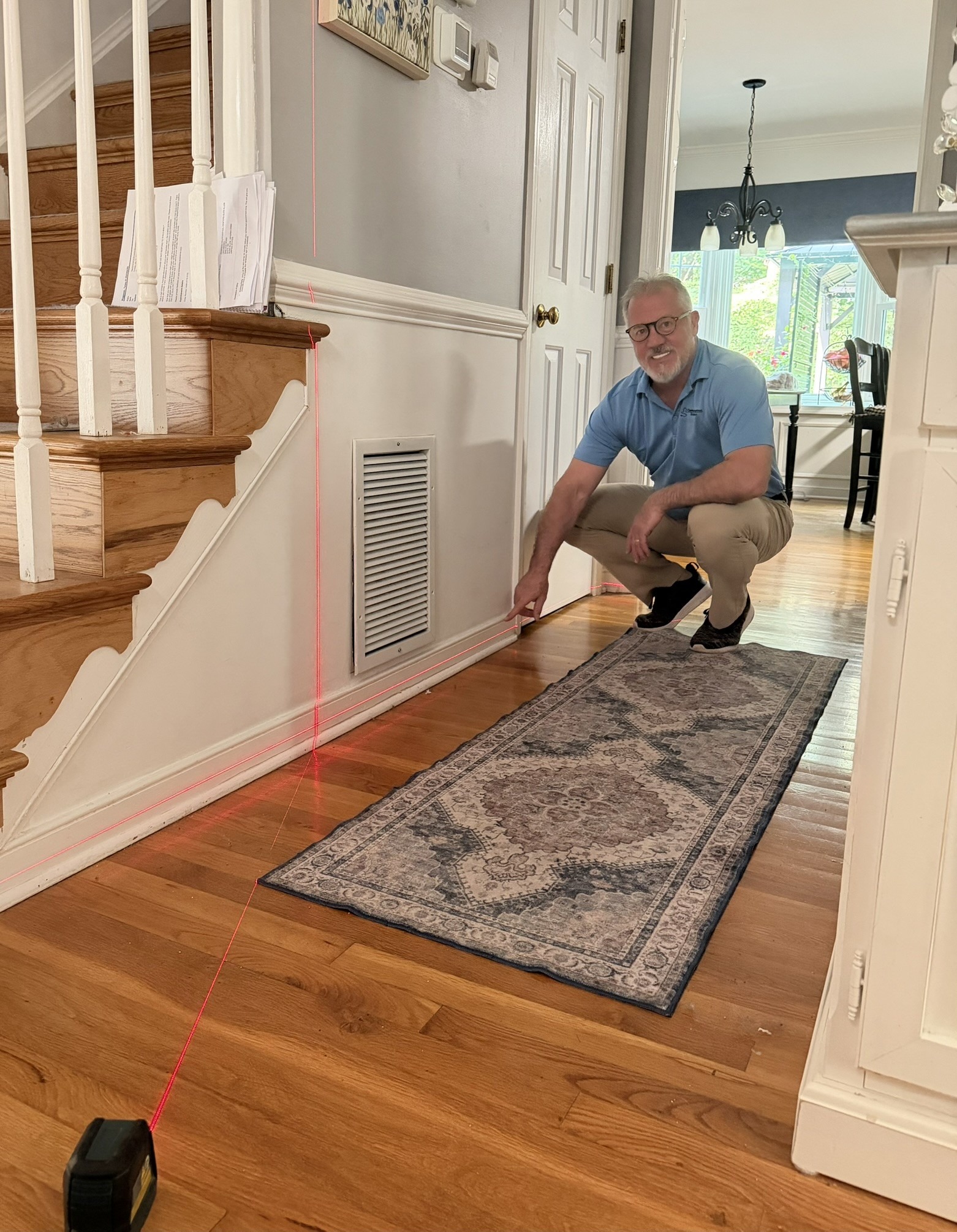 A man kneels beside a red laser line, checking alignment along a hallway with a patterned rug, measuring for home improvement.