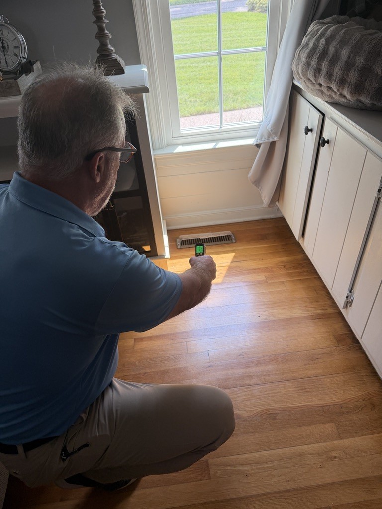 A professional kneels by a vent, using a tool to assess airflow in a well-lit room