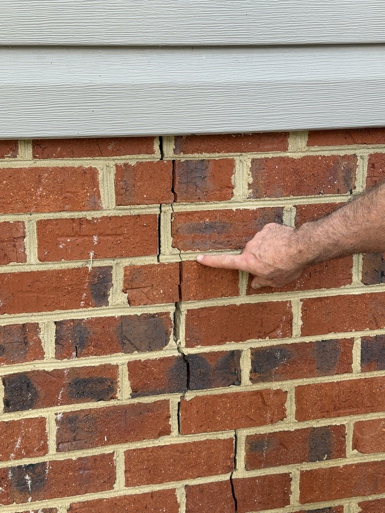 Cracked brick wall with a hand pointing at multiple prominent fissures; highlights potential structural damage and need for inspection.