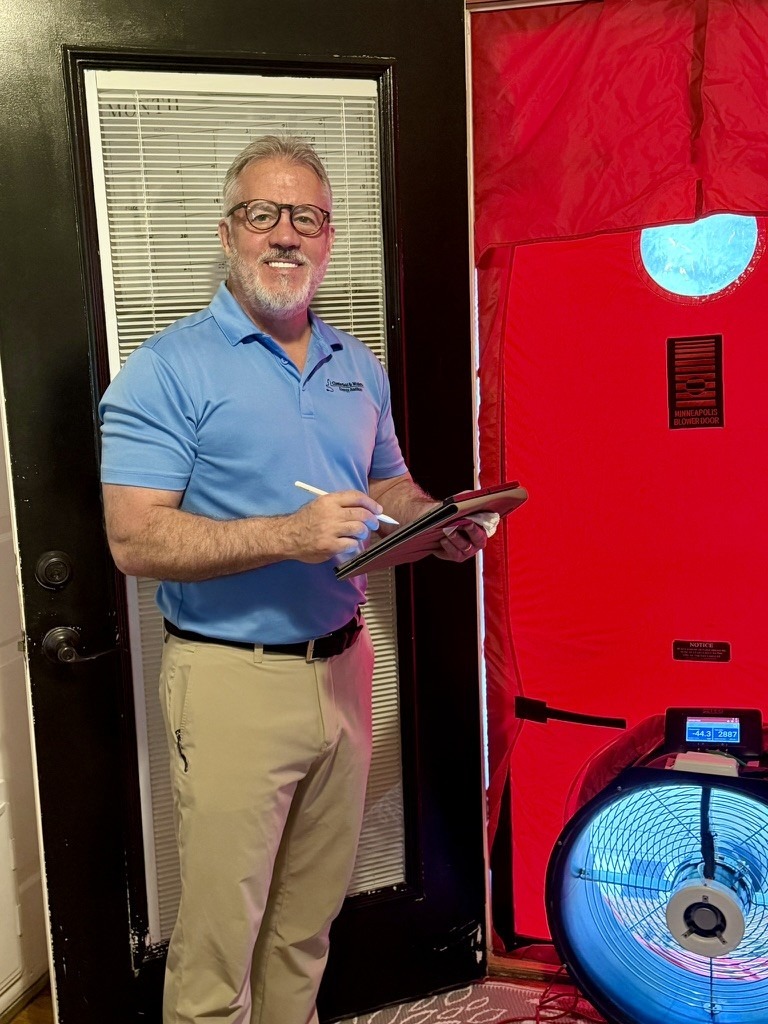 Technician in a blue polo holds a clipboard beside a blower fan and red air pressure testing equipment at a doorway,