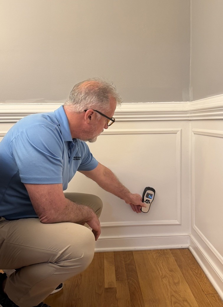 A professional in a blue shirt kneels beside a wall, using a moisture meter to assess the surface's condition, ensuring quality inspection.