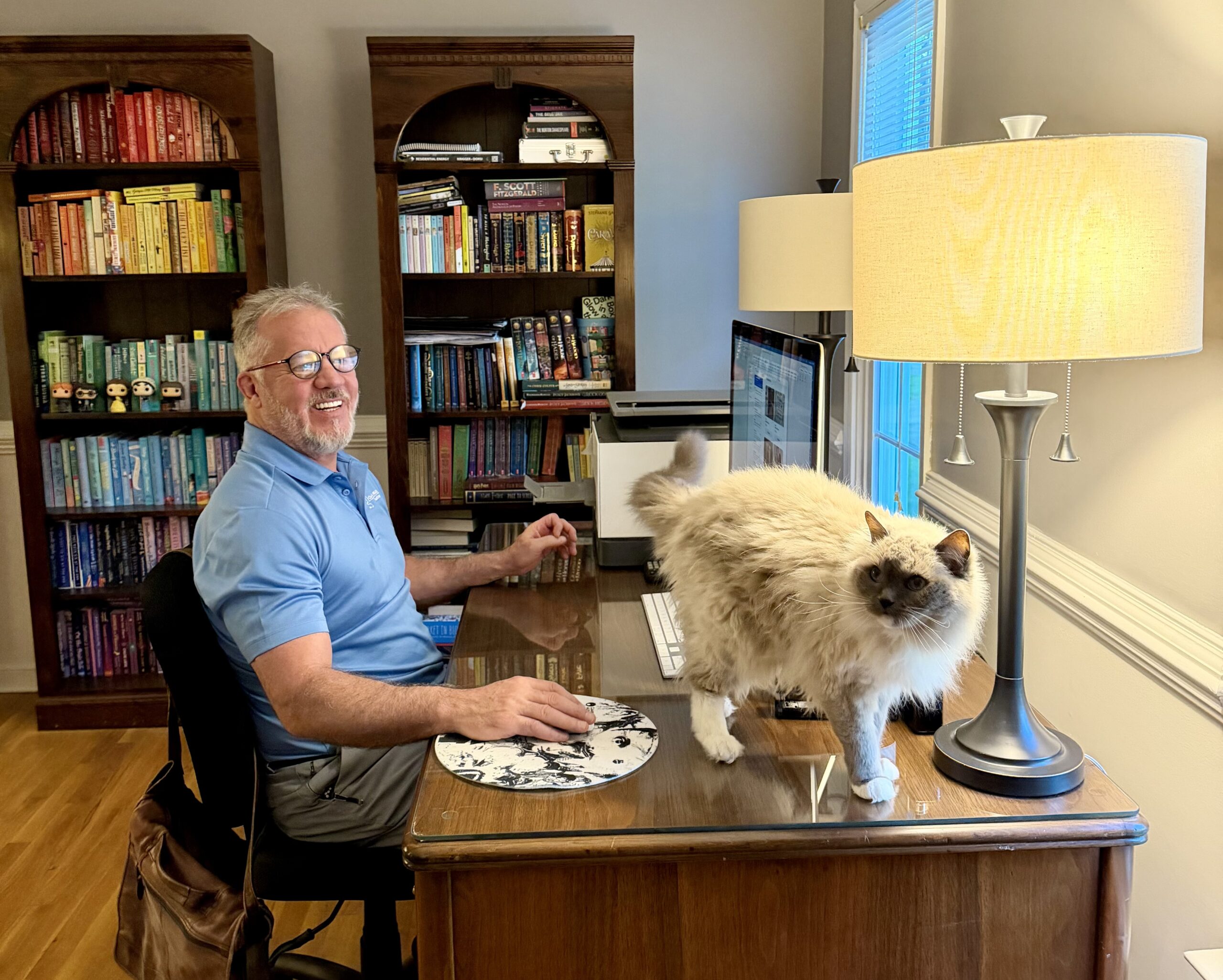 Man smiling at his home office desk with a fluffy cat standing beside his computer.
